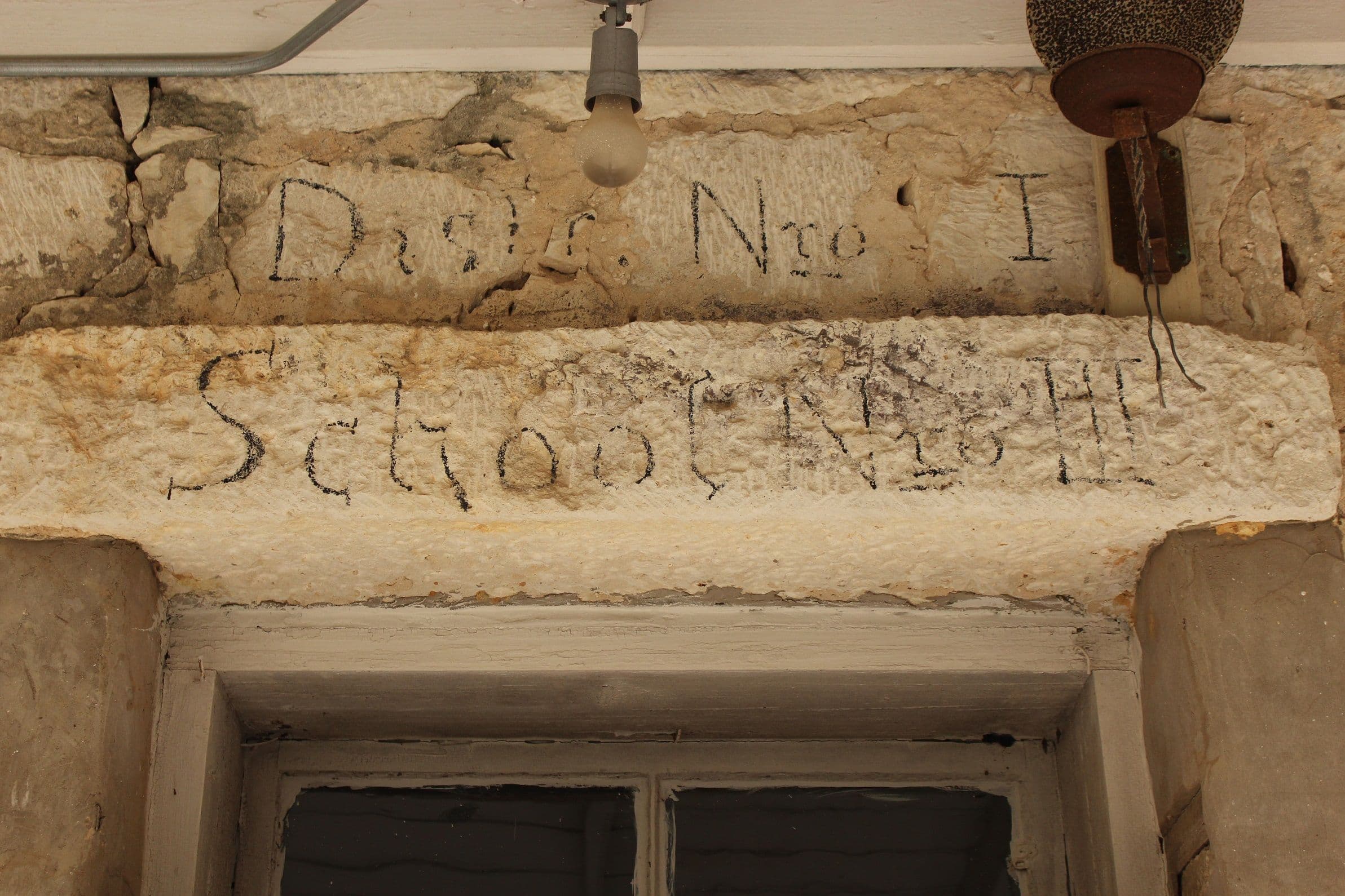Looking up at two rough limestone blocks above a doorway, each carved with darkened hand-cut lettering. A bare light bulb on a porcelain fitting hangs at the left, a rusted lamp shade at the right, and the painted head of a timber door frame is visible below.