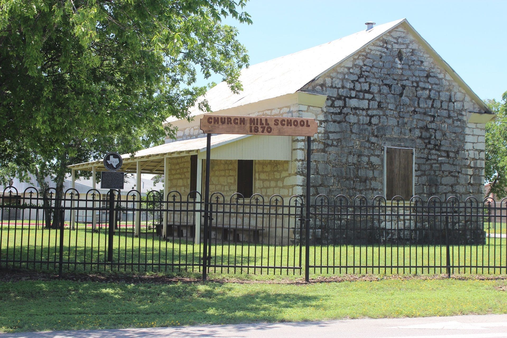 A small single-storey building of rough grey limestone with a metal gable roof and a shed-roofed timber porch along one side, standing behind an ornamental black iron fence, with a wooden sign reading Church Hill School 1870 hung in front.