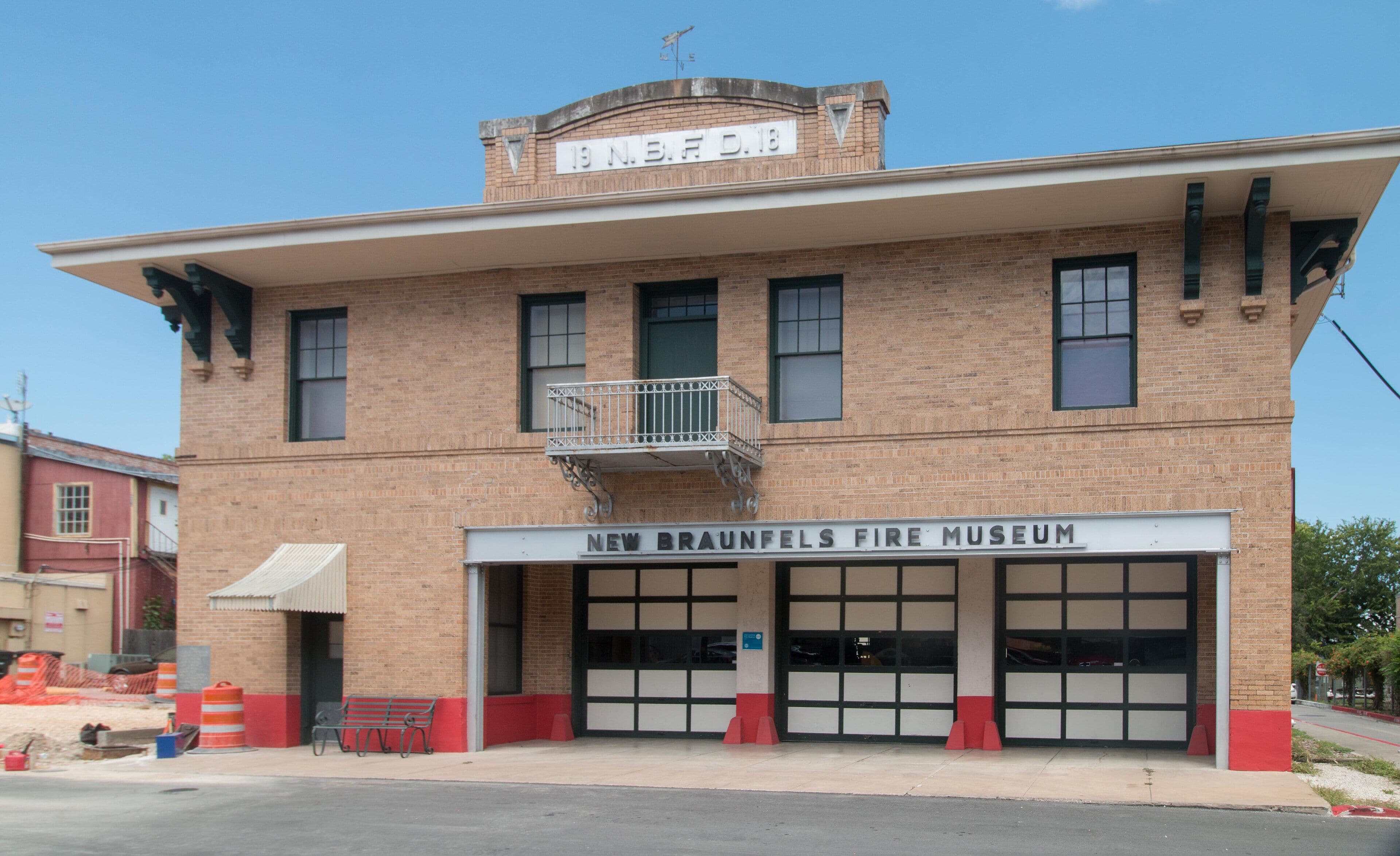 A two-storey buff brick building with a wide overhanging eave on carved brackets. A raised parapet panel at the centre of the roof carries moulded lettering under a weather vane, a small iron balcony projects above, and three wide glazed vehicle doors sit below a sign reading New Braunfels Fire Museum.