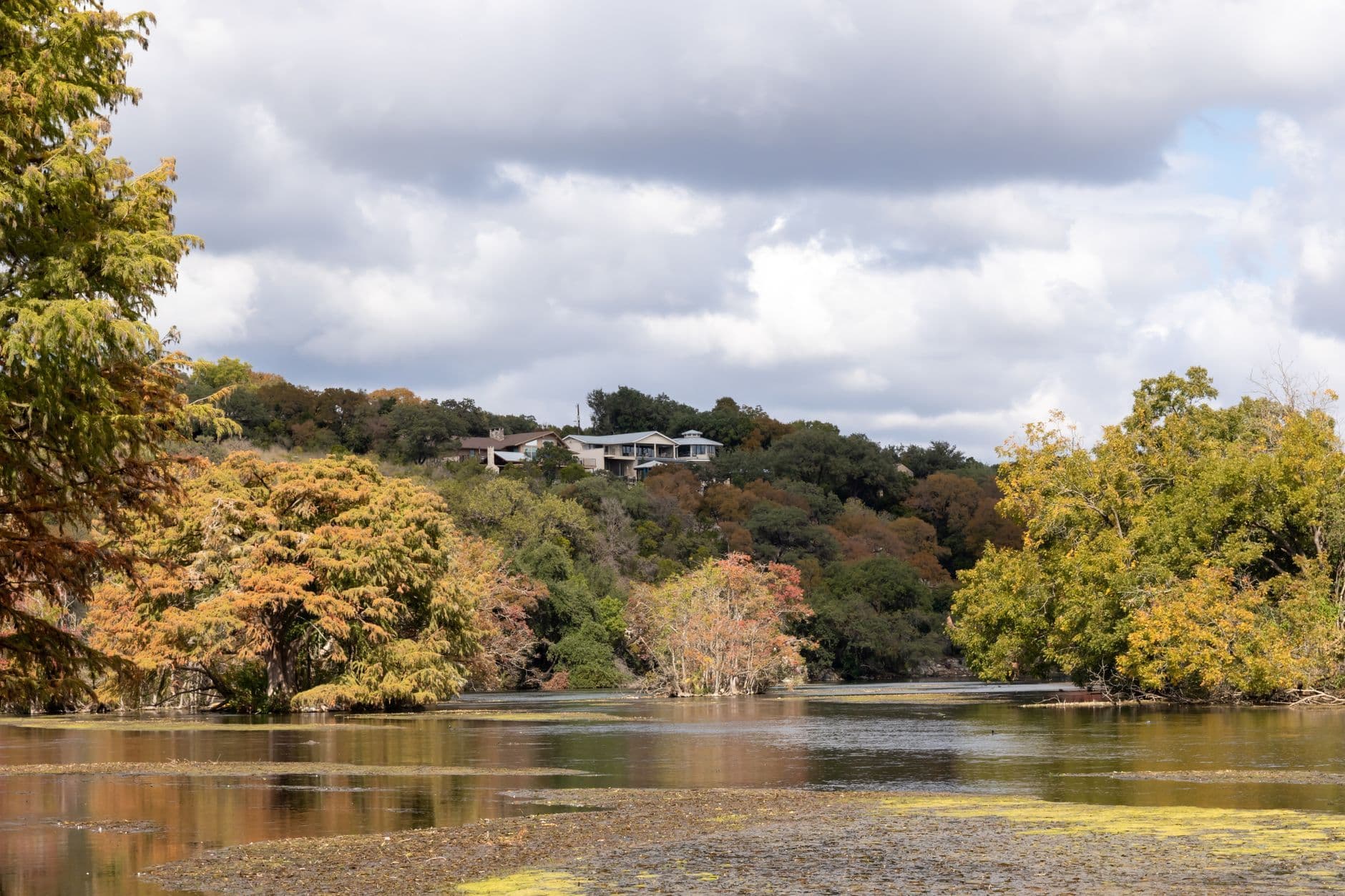 Wide view across a river toward autumn trees and distant hillside homes under a cloudy sky.