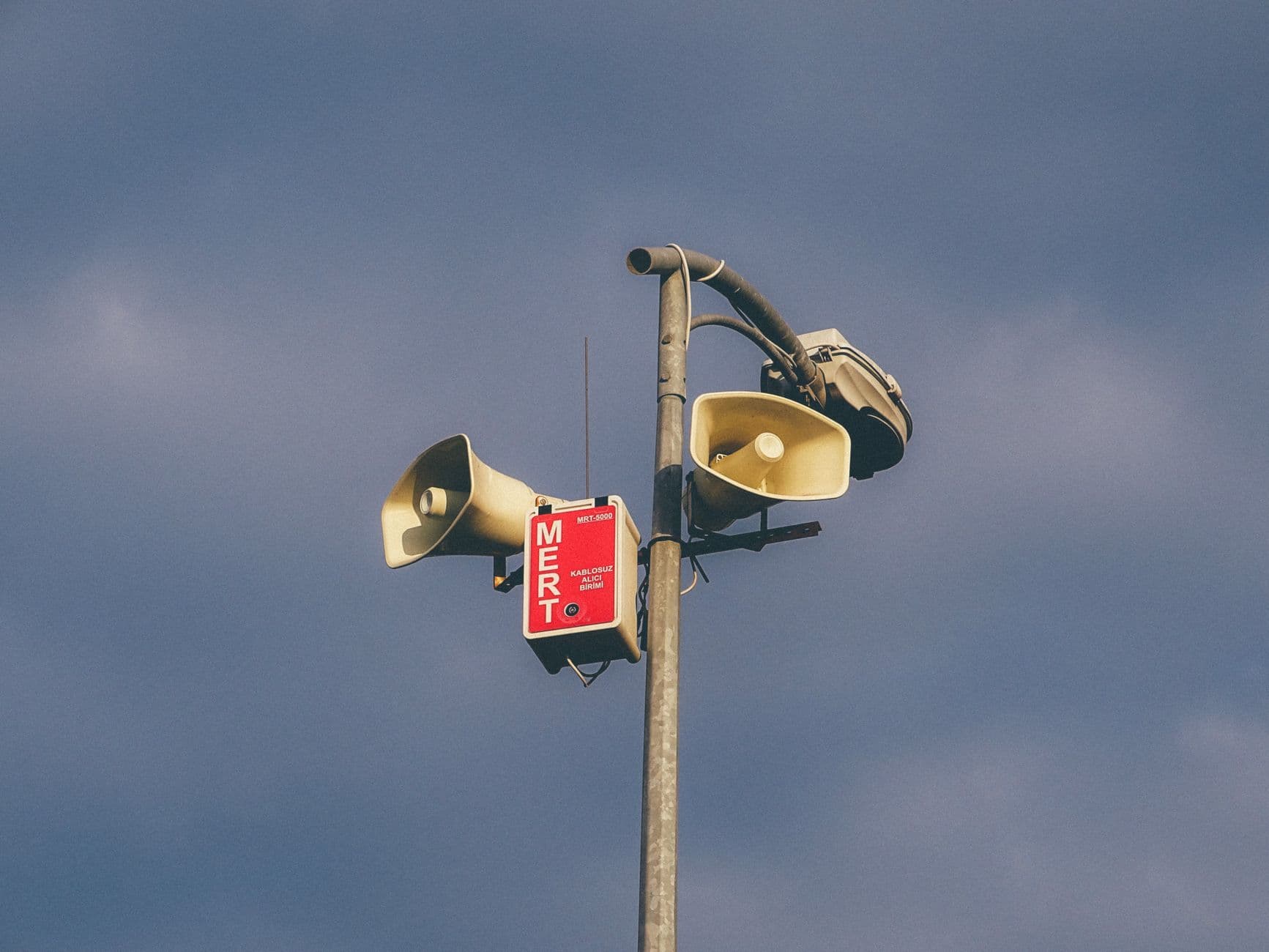 Several horn loudspeakers and a red equipment box mounted on a metal pole against the sky.