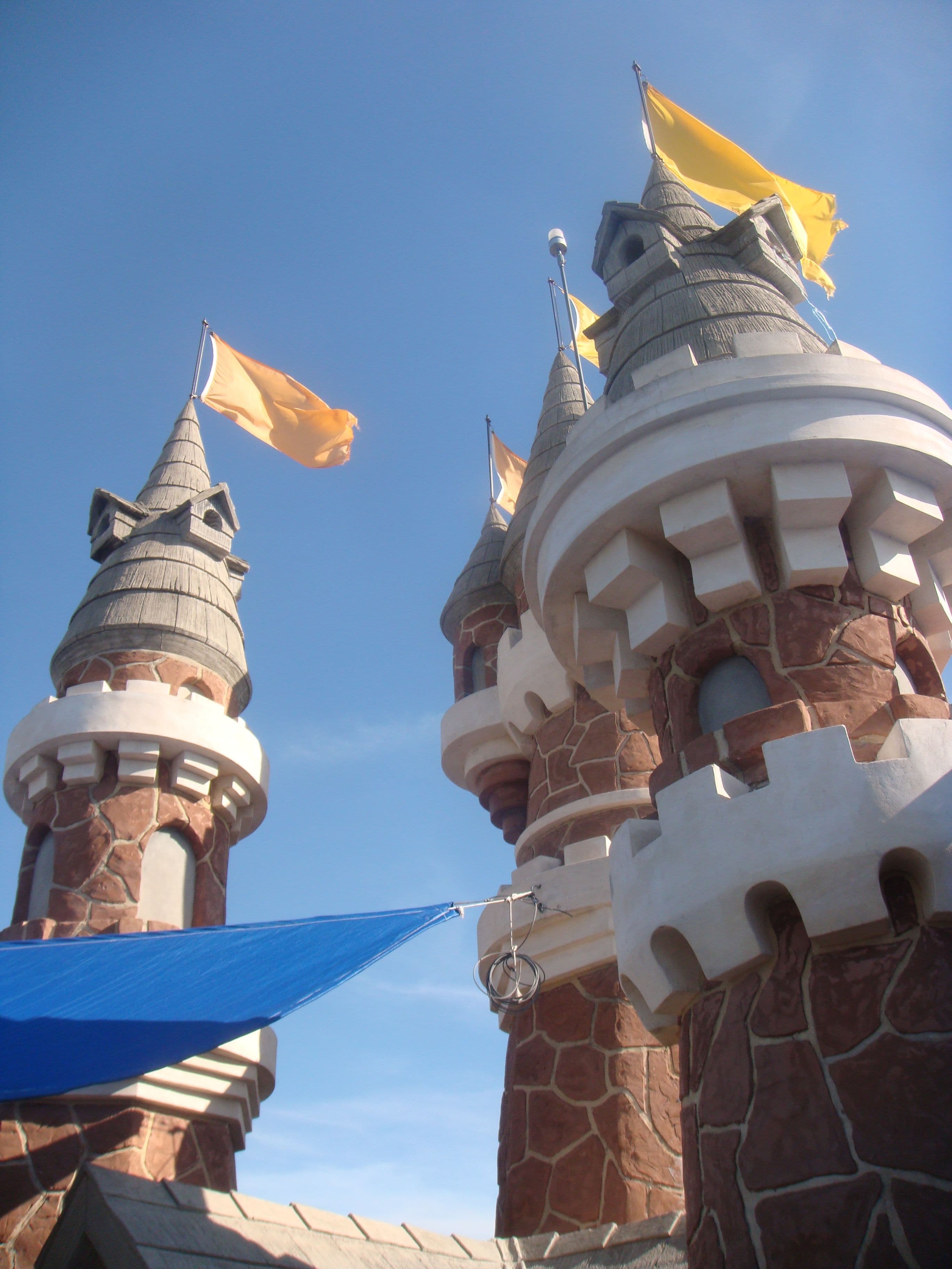 Three castle-style turrets with battlemented tops and conical grey roofs rise against a deep blue sky, their walls faced in irregular red-brown stone with cream trim. Orange and yellow pennants fly from staffs at the tops, and a blue triangular shade sail is tensioned across the lower left.