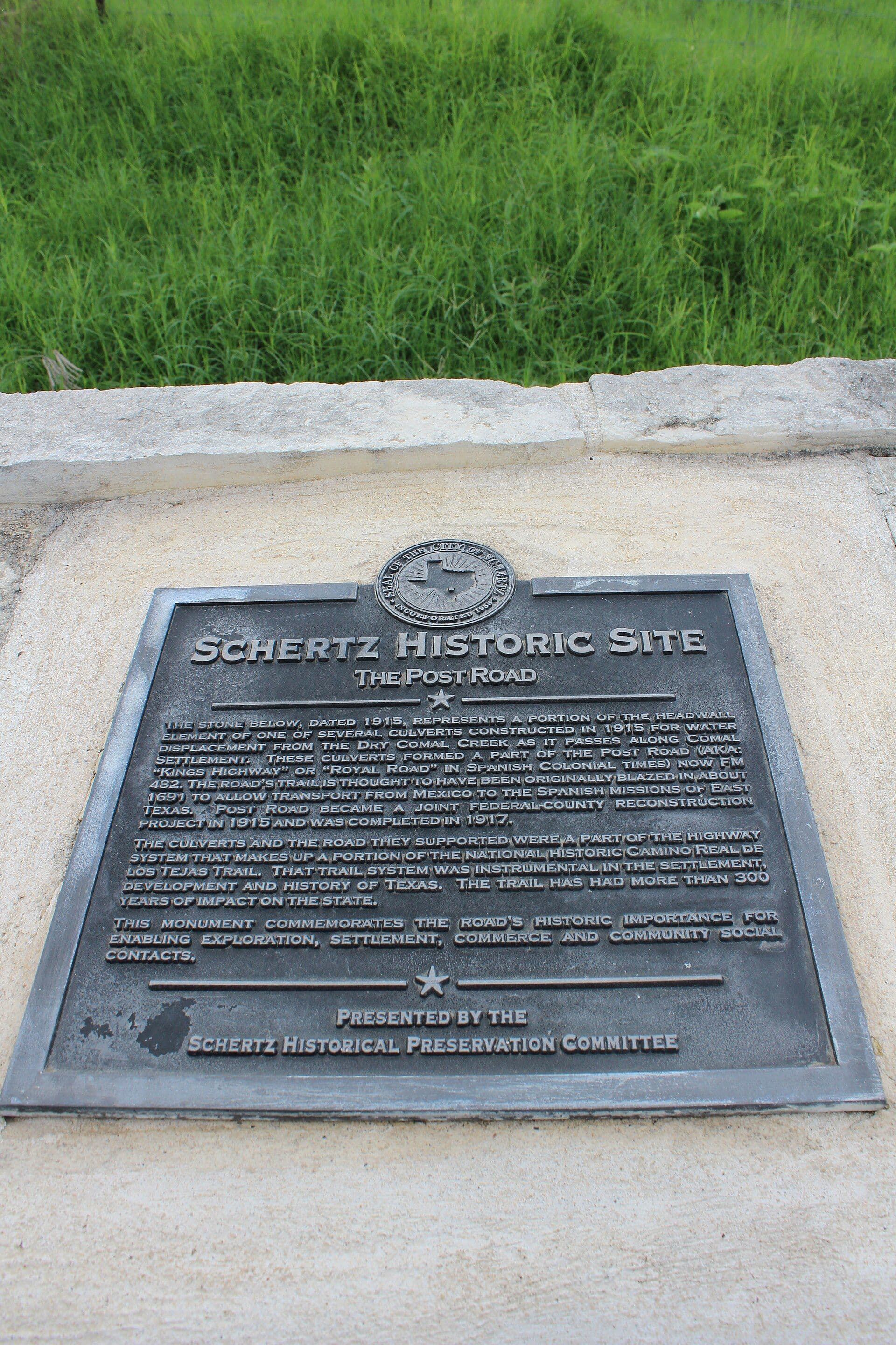 A dark cast-metal plaque set flush into the pale sloping face of a low masonry monument, headed by a round city seal with an outline of Texas and reading 'Schertz Historic Site — The Post Road' above three paragraphs of raised text. A rough limestone block edges the monument behind, with tall green grass beyond.
