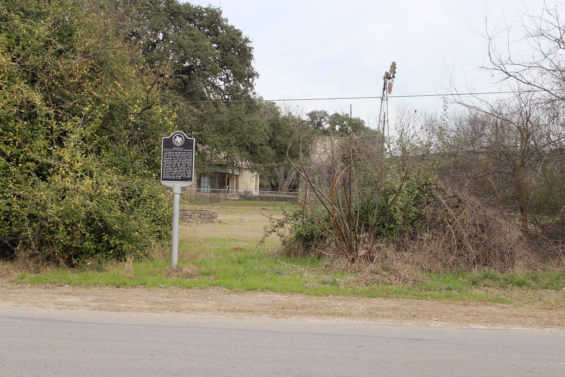 A black Texas historical marker with silver lettering on a grey post stands in the grass beside a narrow asphalt road. Behind it an open mown lot leads to a small stone building with a metal awning behind a chain-link fence, framed by heavy brush at left and a windmill tower at right under a white sky.