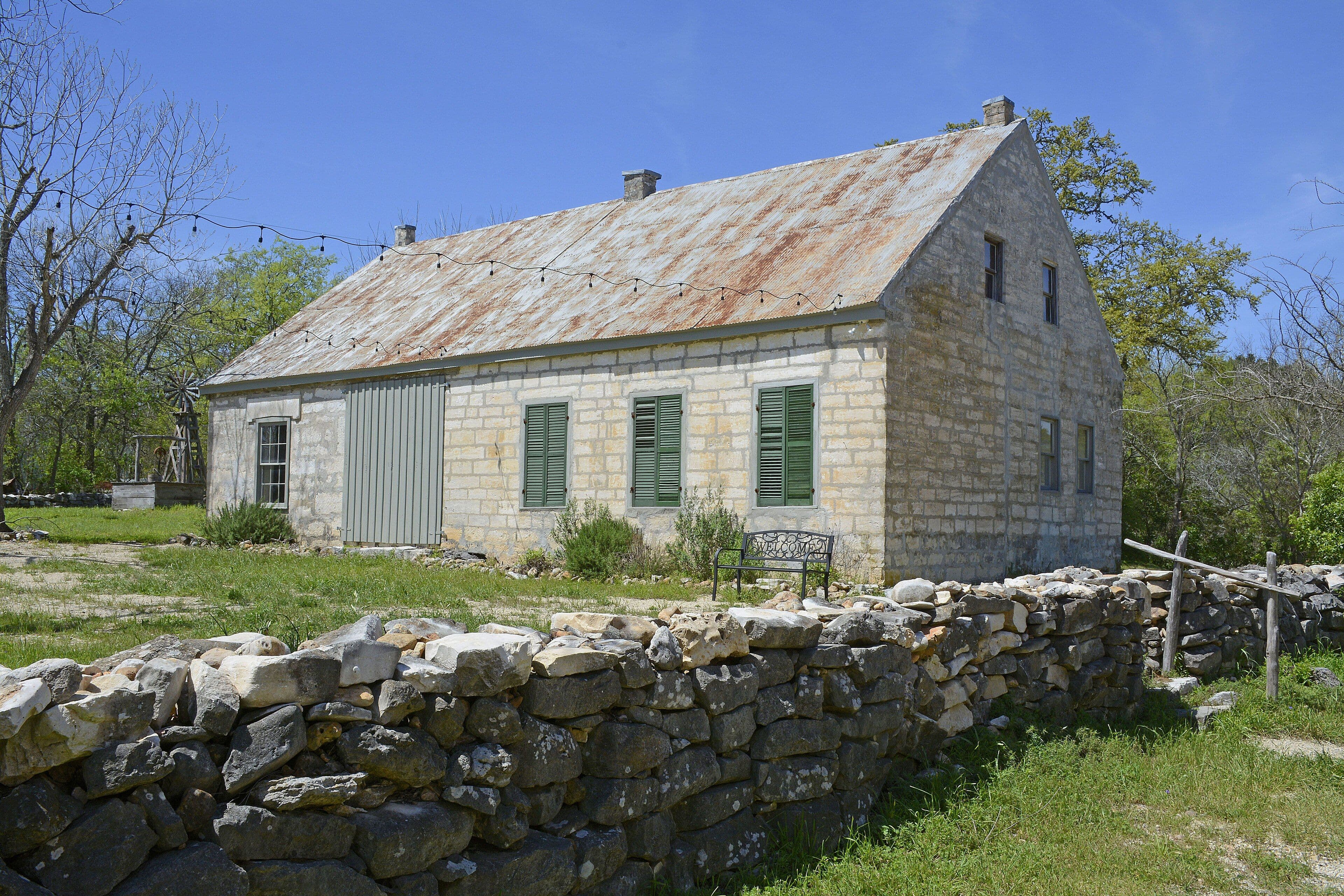 A long, low limestone farmhouse with a steep rust-streaked metal roof and green louvered shutters, standing behind a waist-high wall of dry-stacked limestone. A windmill tower and oak trees are behind it under a deep blue sky.