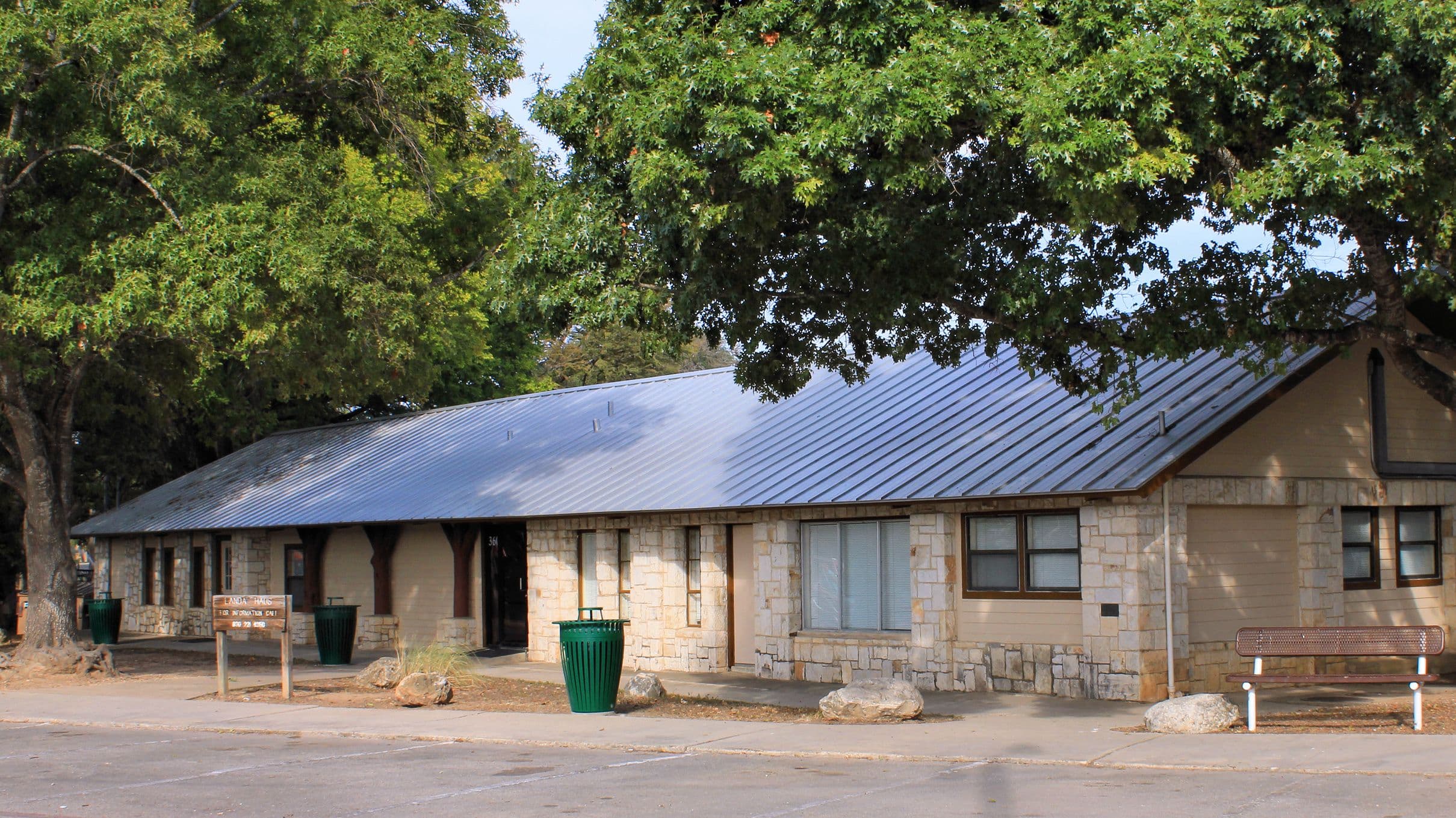 A long single-storey park building with cream cut-limestone walls, stucco panels and a broad grey metal hipped roof, seen at an angle across an empty asphalt lot. Green litter bins, a metal bench and a low timber sign stand along the front, shaded by large oaks.