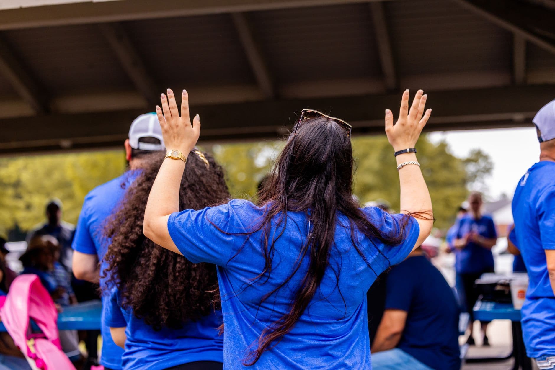 Rear view of a person in a blue shirt raising both hands amid a group under an outdoor shelter.