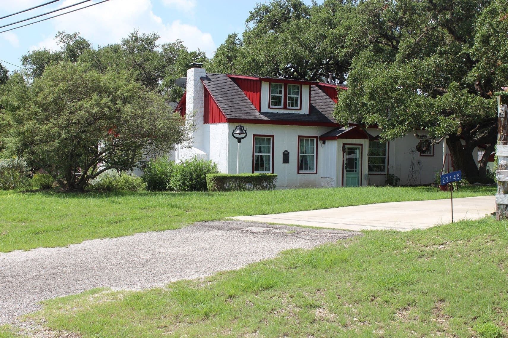 A white-painted masonry building with dark red window trim, a red metal roof slope and a three-window dormer, shaded by live oaks, with a large iron bell on a post in the front yard.