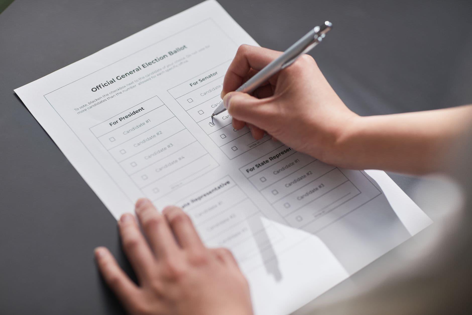 Hands marking a checkbox on a printed election ballot with a silver pen.