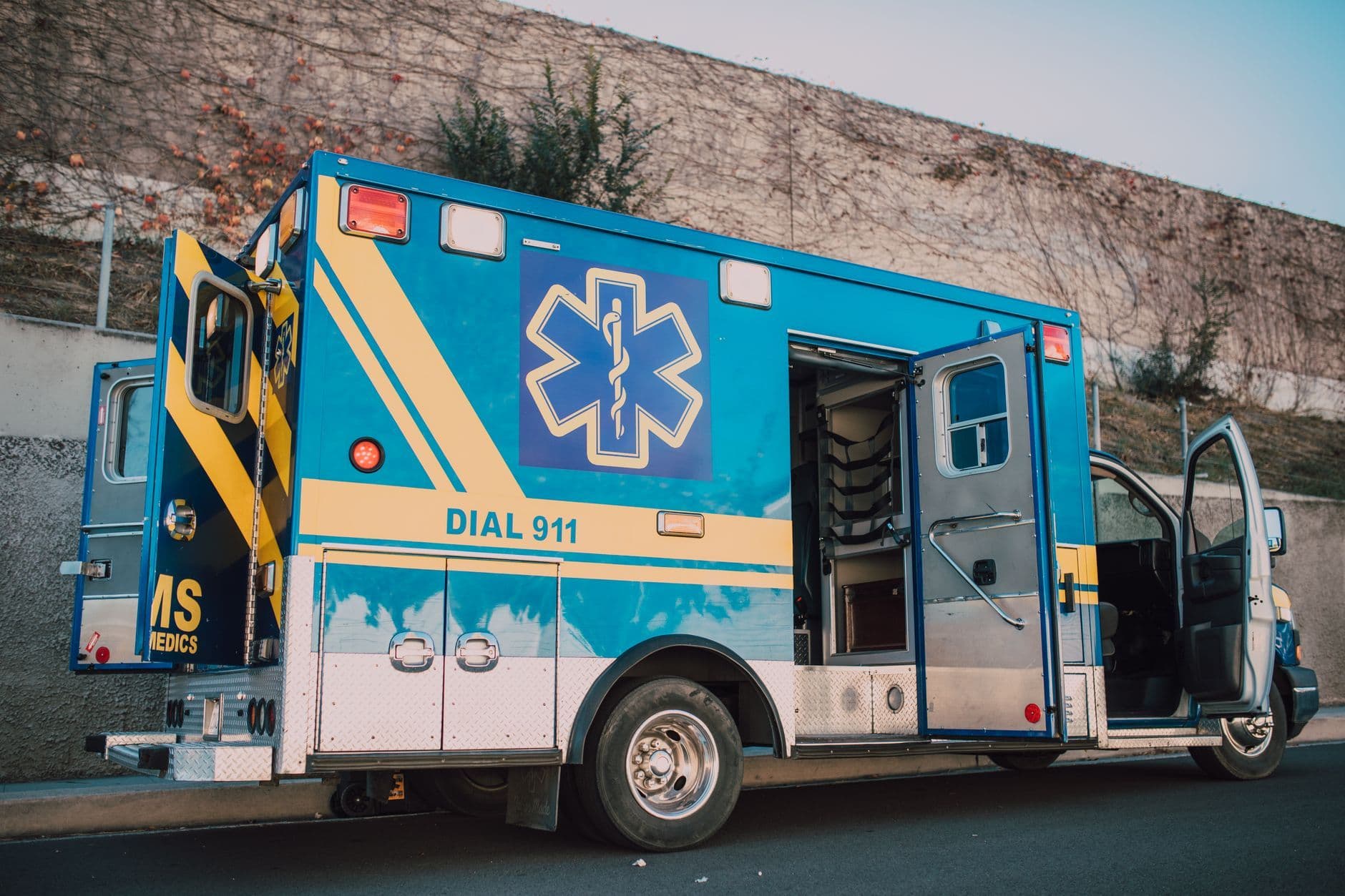 Blue and yellow ambulance parked at a curb with open doors and an emergency medical symbol on its side.