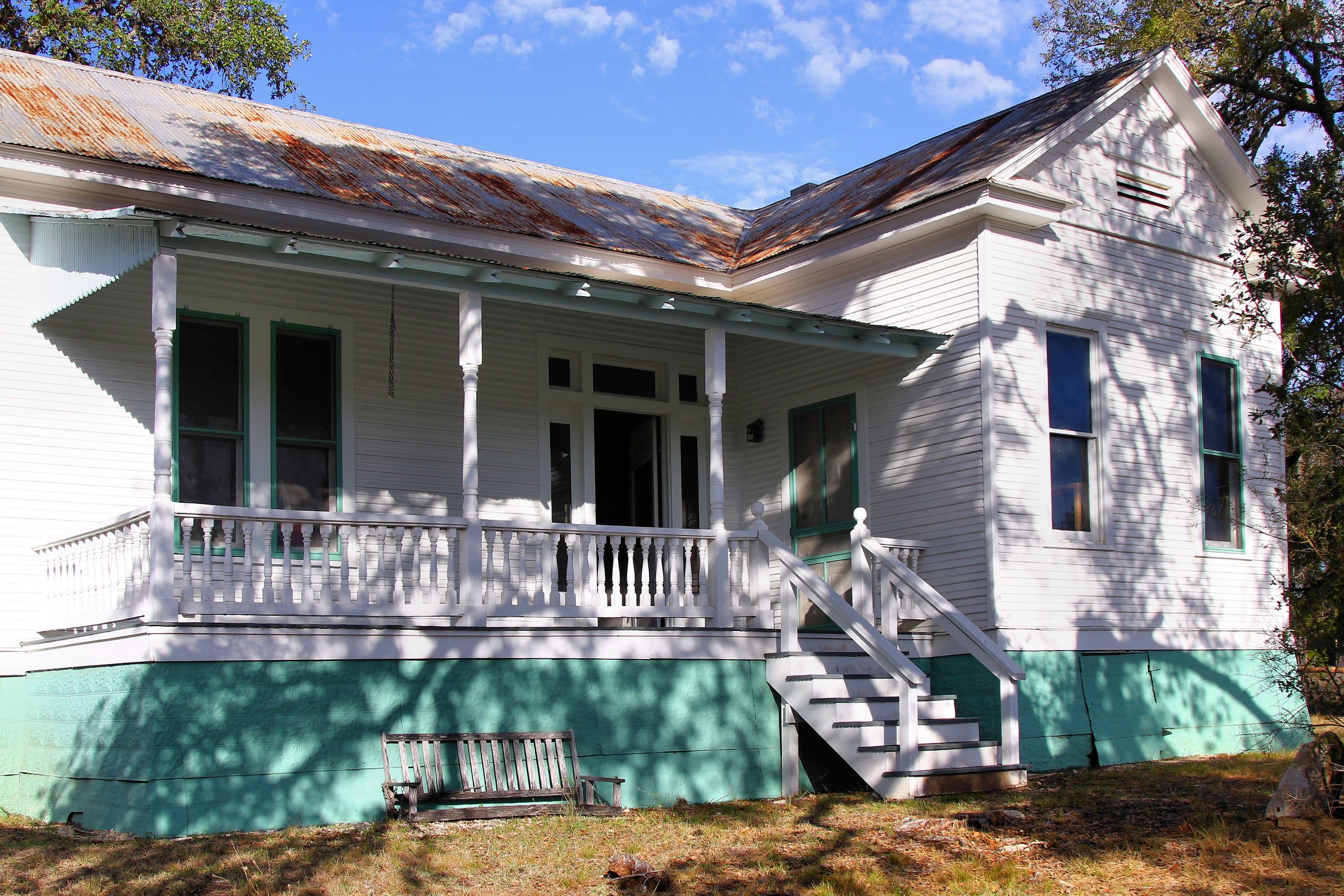 A one-storey white wooden house with a deep porch behind turned posts and a spindle railing, teal-trimmed windows, a mint-green painted foundation skirt and a heavily rusted corrugated metal roof, framed by live oaks under a blue sky.