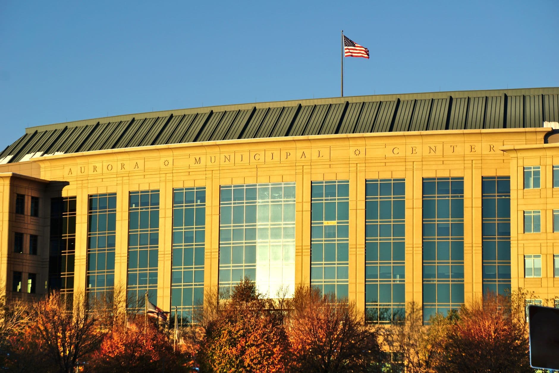 Sunlit municipal center exterior with tall windows, autumn trees and a U.S. flag against a clear blue sky.