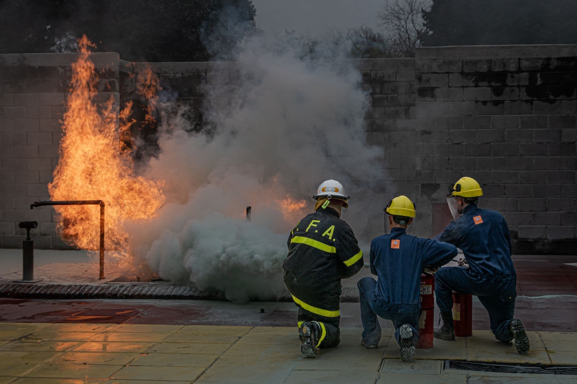Three helmeted trainees kneeling with fire extinguishers near flames and thick smoke in an outdoor training area.