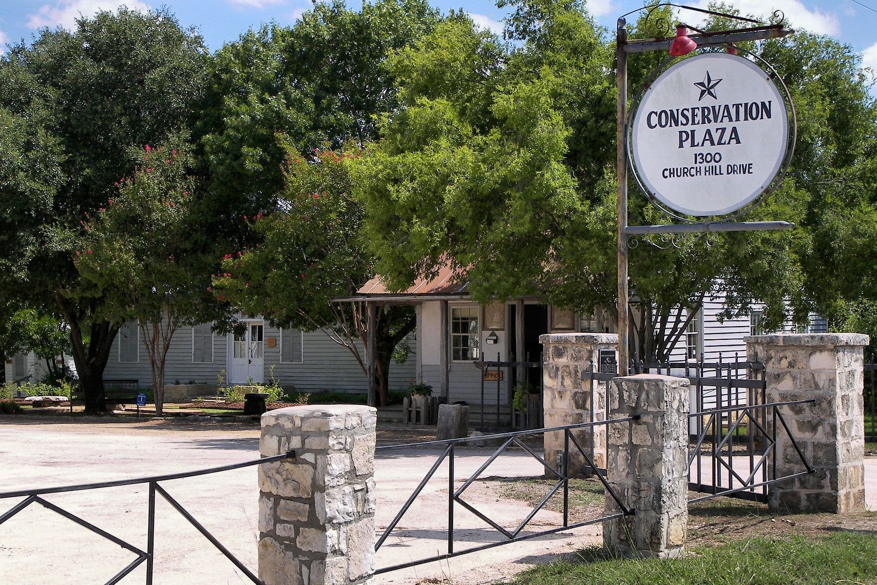 A round white sign lettered CONSERVATION PLAZA hangs from a metal post beside an open gate. Beyond it, limestone piers and a black rail fence front a gravel drive leading to a white wooden building with a rusty metal roof, shaded by large trees.