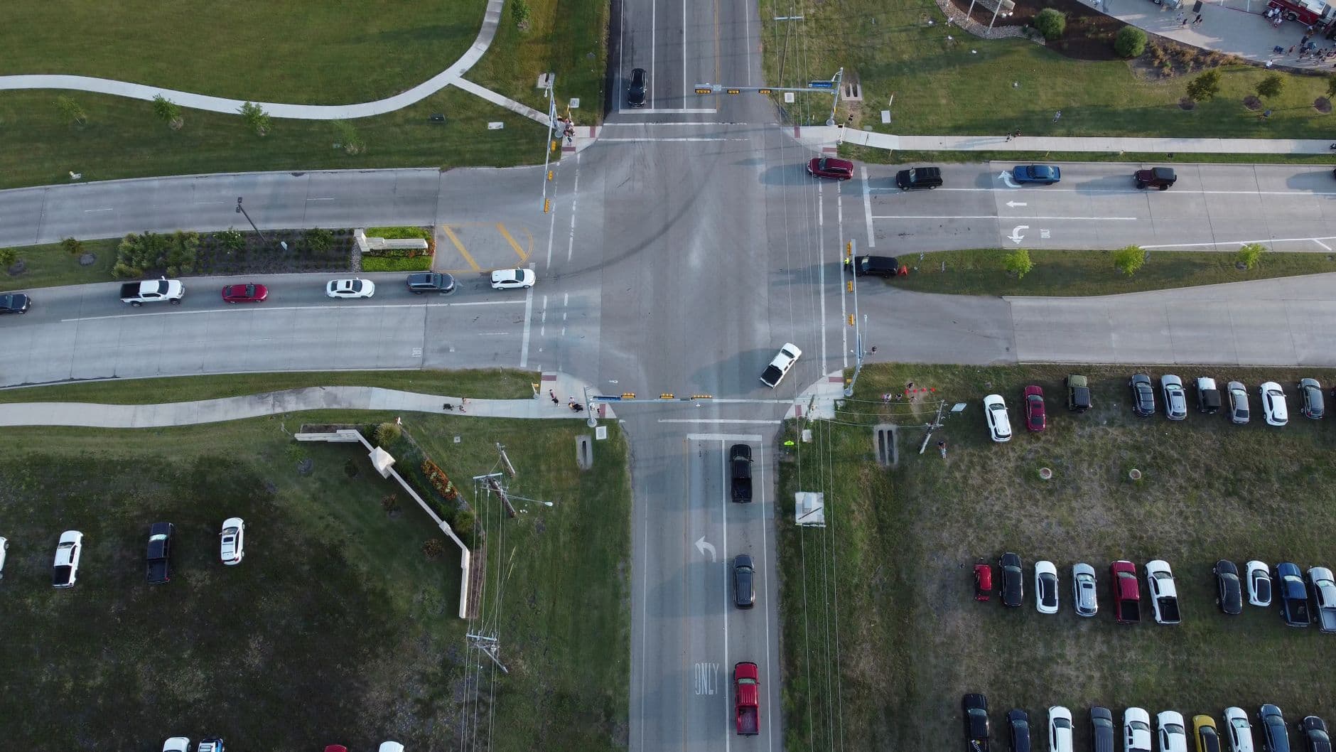 Overhead view of a wide signalized road intersection bordered by grass, sidewalks, utility lines and parked vehicles.
