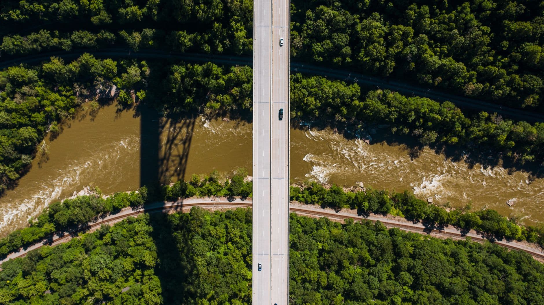 Aerial view of vehicles on a divided bridge crossing a river, with railroad tracks and dense green trees below.