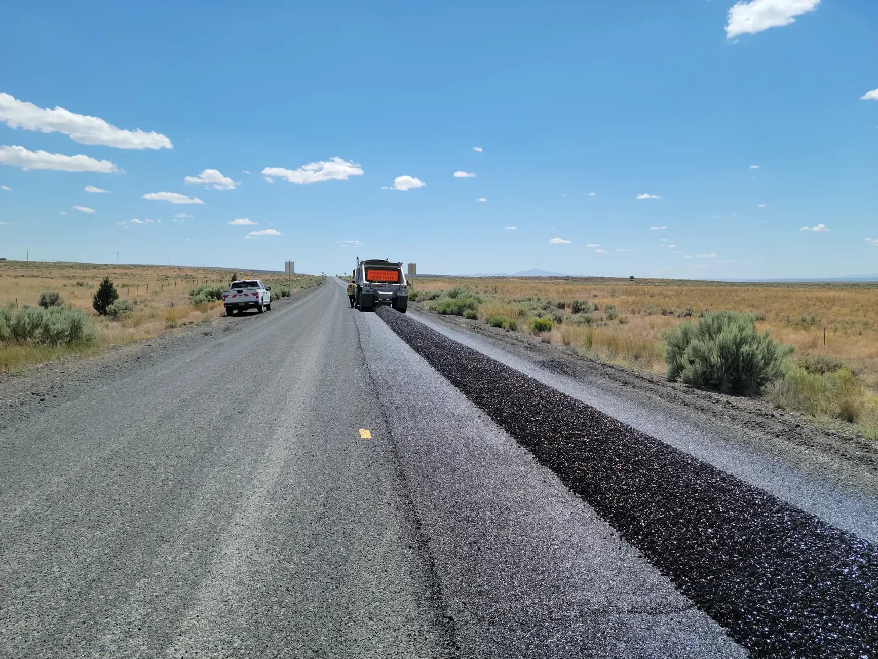 Roadwork vehicle laying fresh asphalt on a rural road beneath a blue sky.