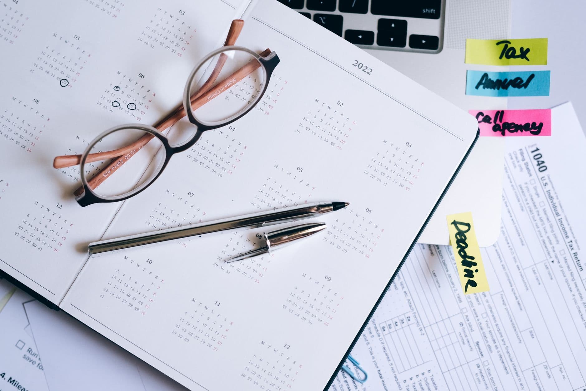 Open 2022 calendar with eyeglasses, pens, tax forms and colorful reminder notes on a desk.