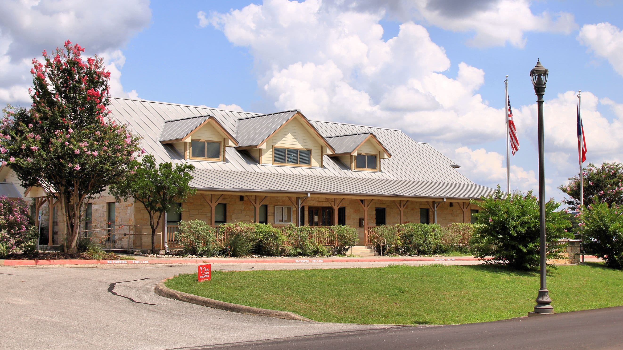 A long single-storey civic building with tan limestone walls, cream siding and a grey standing-seam metal roof with three dormers. A timber-posted porch runs along the front, American and Texas flags fly on poles to the right, and a pink-flowering crape myrtle stands at the left above a mown lawn.