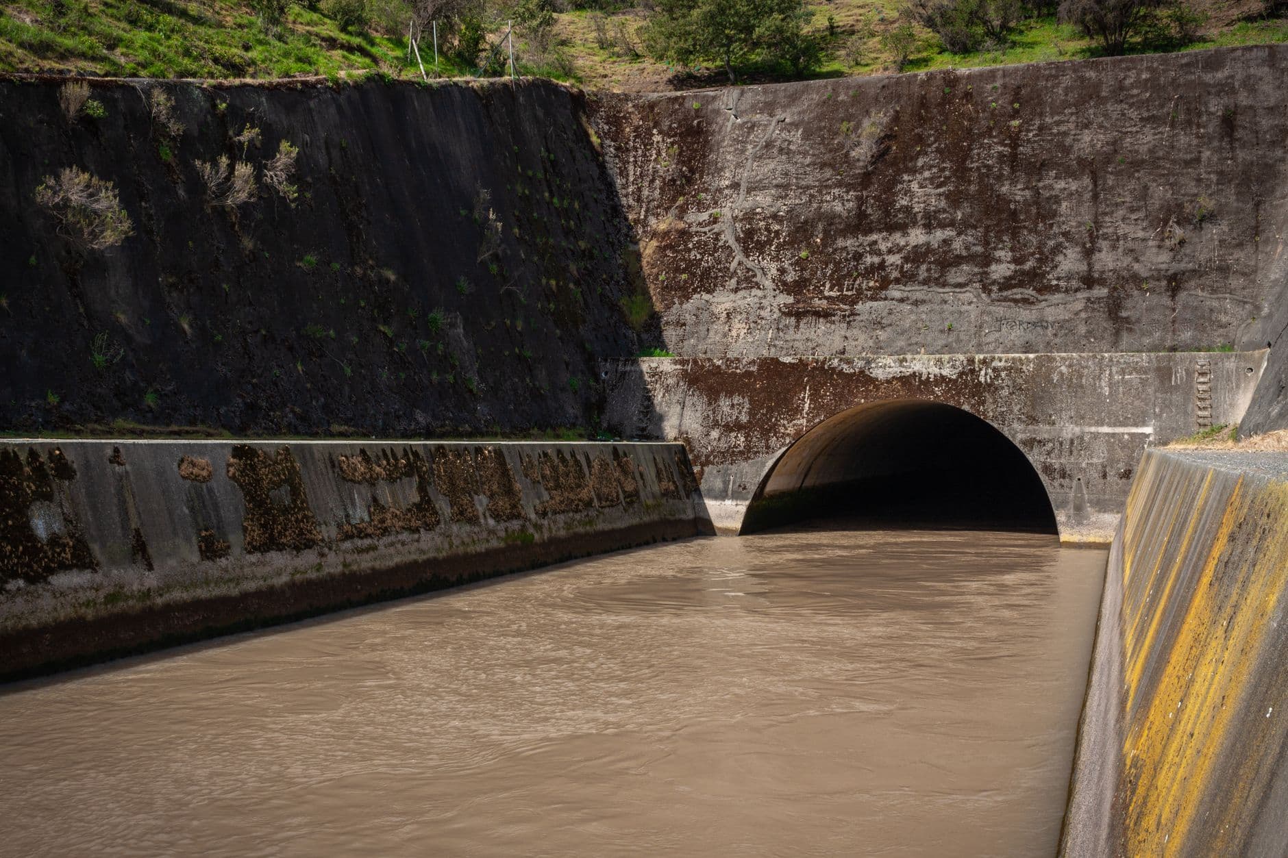 Muddy water flowing through a concrete-lined drainage channel toward a large arched culvert.