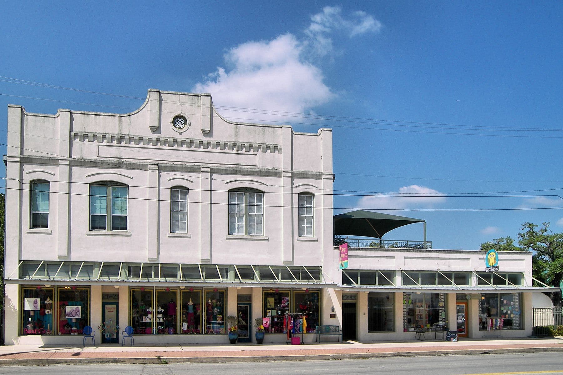 A two-storey off-white rendered commercial building with a curved decorative parapet and five arched upper windows, seen straight on from across the street. Metal awnings shade the shopfronts below, and a lower single-storey shop range adjoins it on the right.
