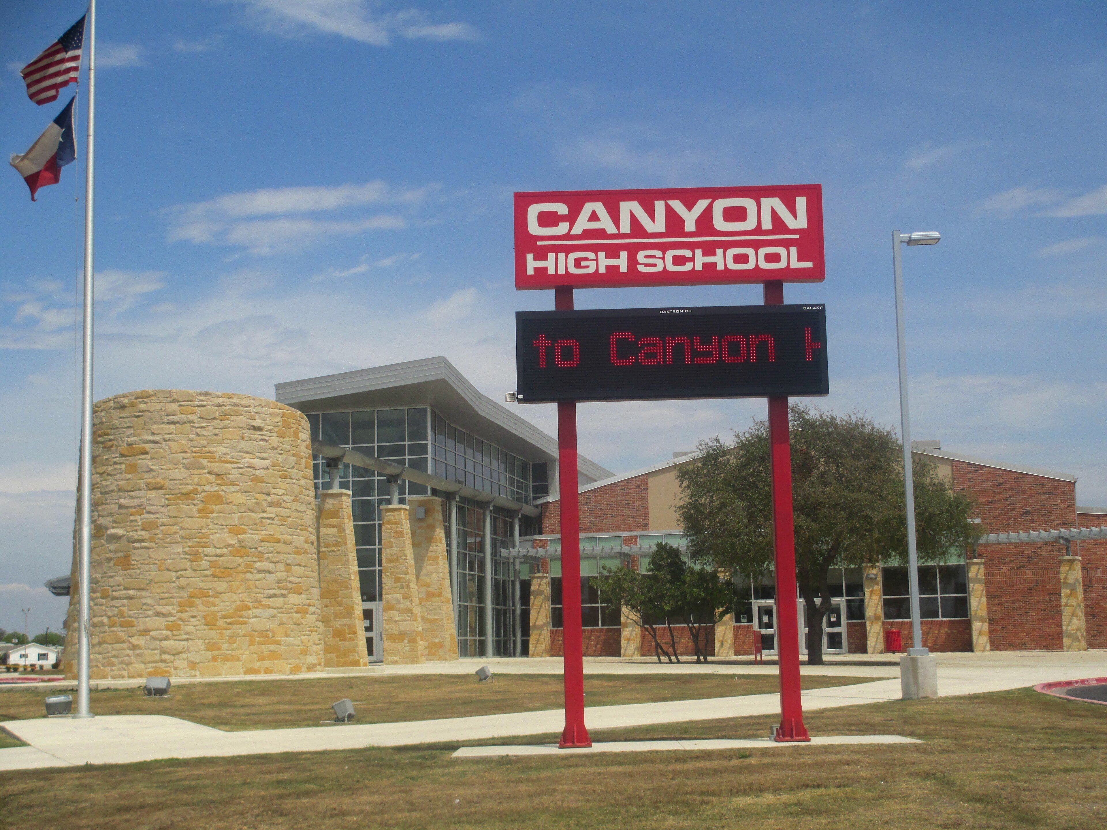 A tall red sign reading Canyon High School stands above an electronic message board on a mown lawn. Behind it are a curved stone drum, a glass-walled entrance under a curved metal canopy, and long brick classroom wings. A flagpole flies the United States and Texas flags at the left.