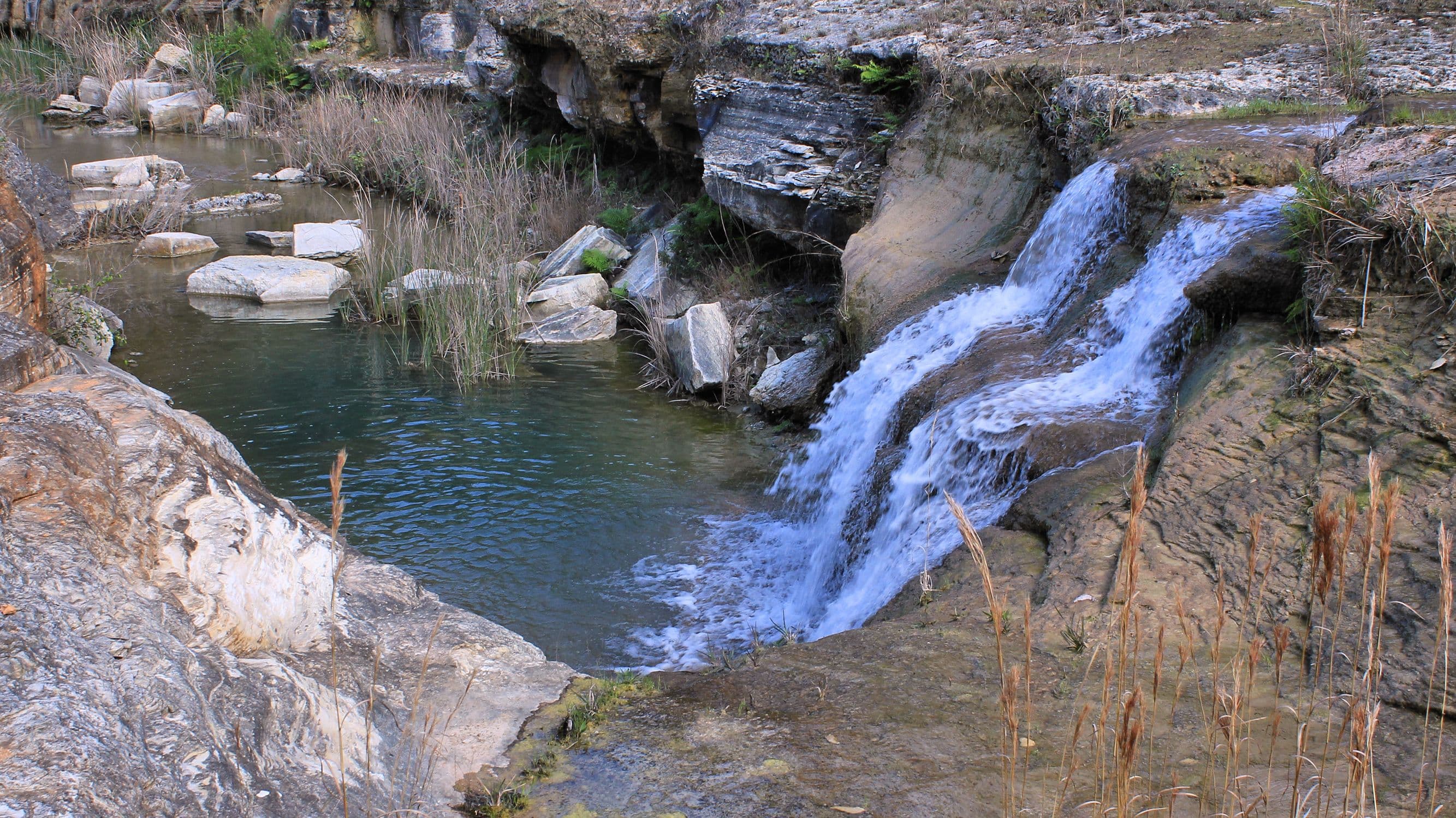 A wide, shallow waterfall pours over stepped grey limestone ledges into a dark green pool. Bare rock, angular boulders and dry grass line the banks, with a rocky channel running off in the background.