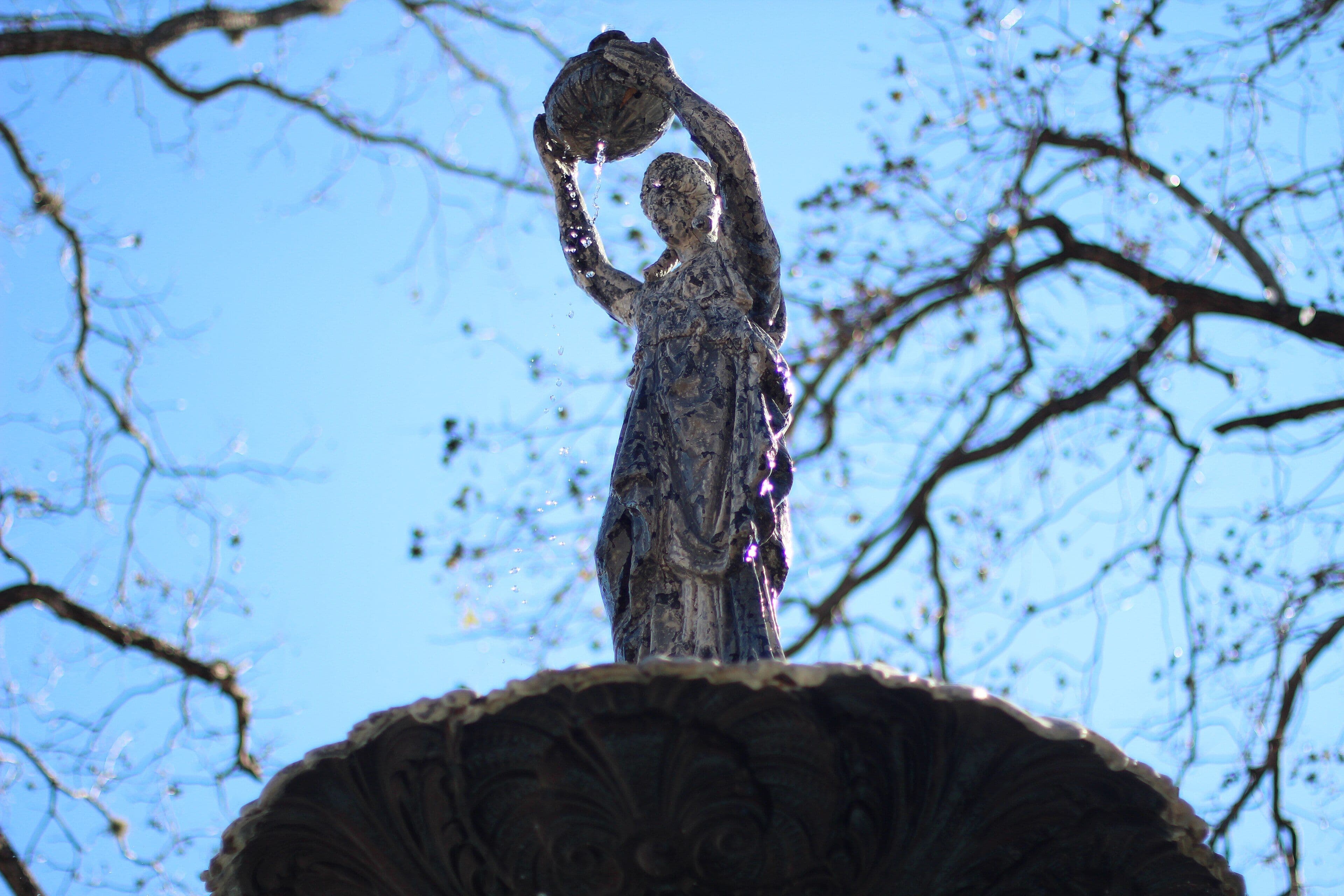 Looking up at a weathered metal statue of a draped woman holding a shallow bowl above her head, with water spilling from it. She stands on the ornate top bowl of a fountain, against a blue sky crossed by bare branches.