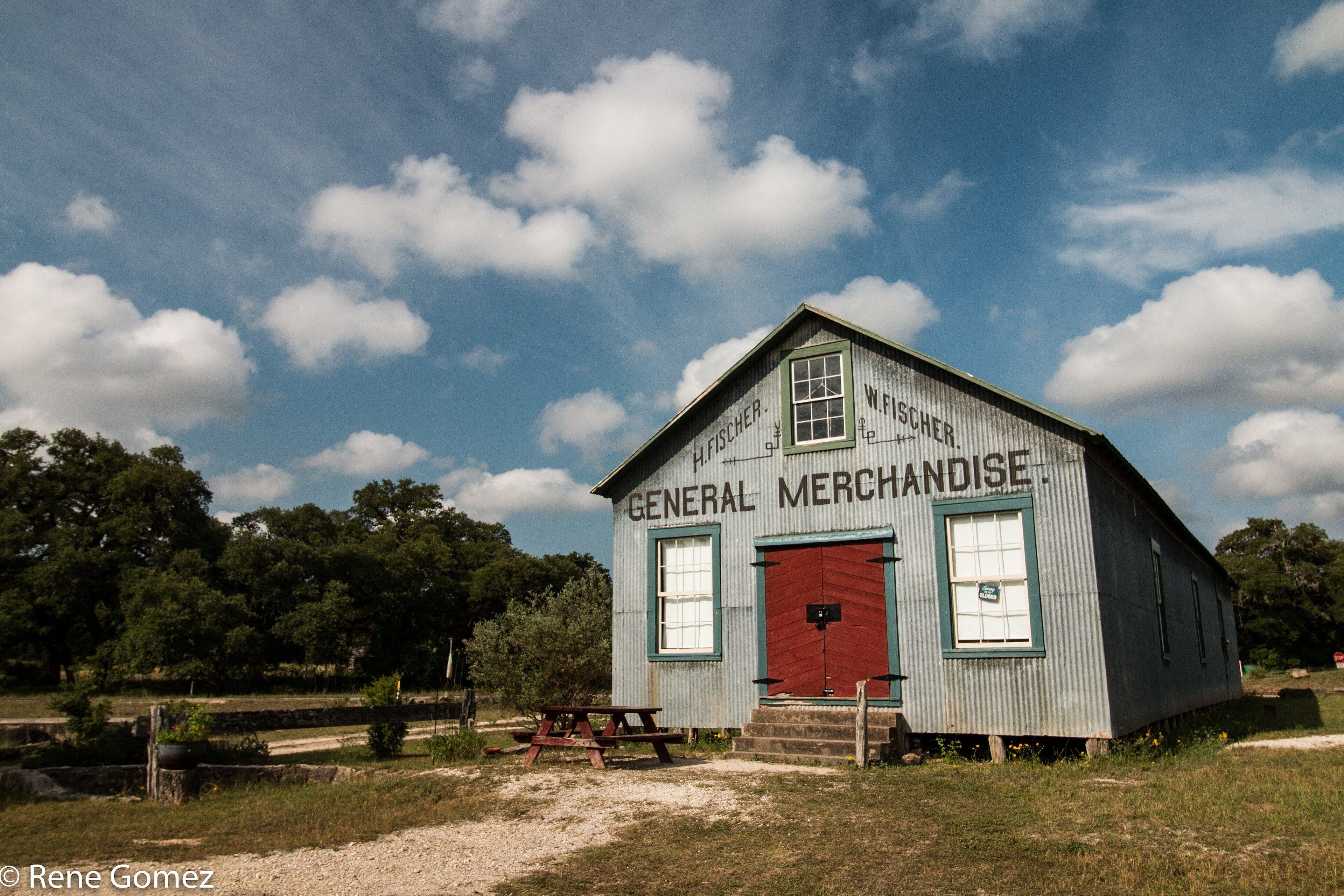 A long single-storey building clad in corrugated grey metal with a red double door and green-framed windows. Painted black lettering across the gable reads GENERAL MERCHANDISE. Gravel paths, a picnic table and oak trees stand in front under a cloudy blue sky.