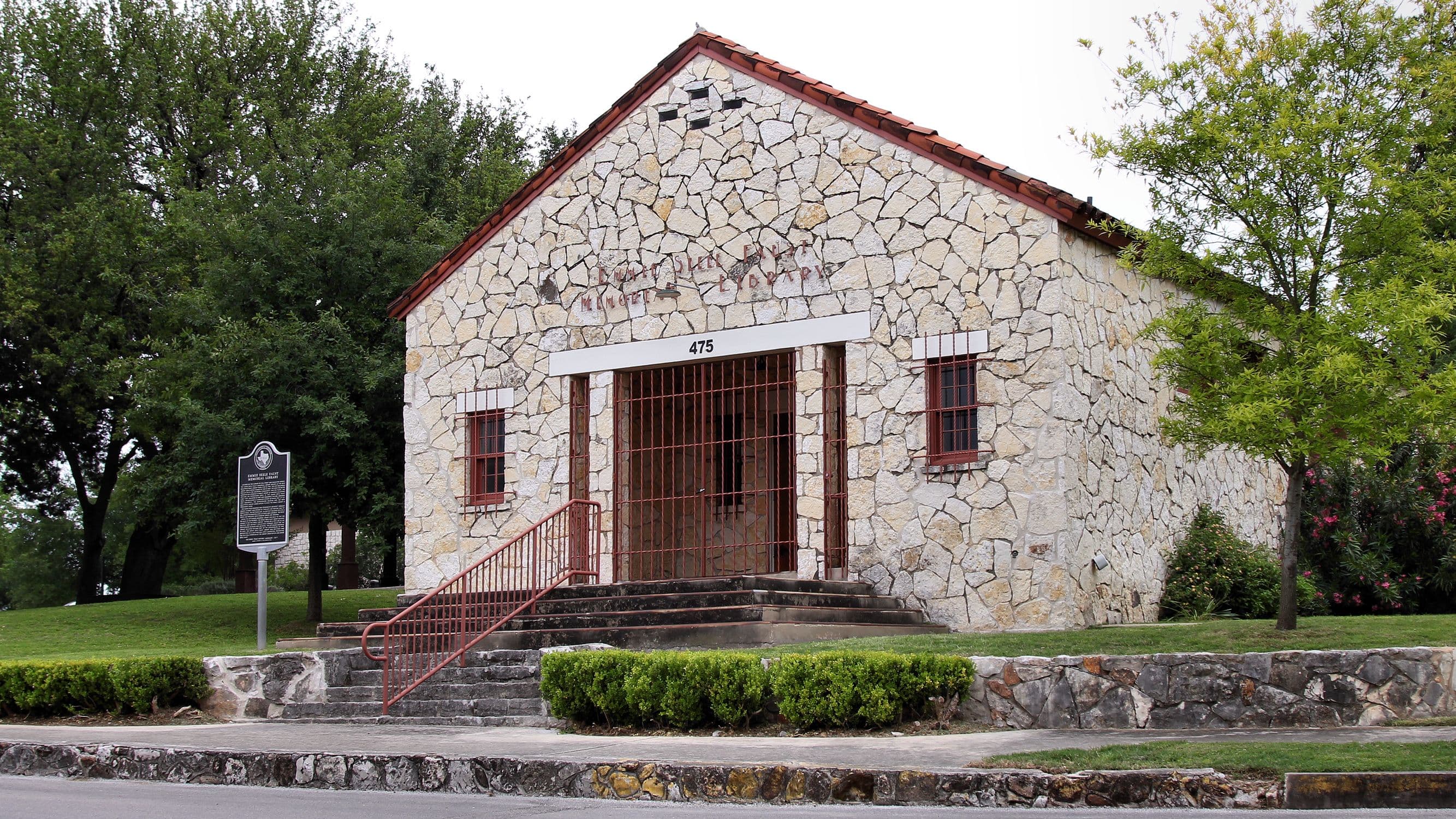 A one-storey building of irregular cream limestone with a red barrel-tile gable roof sits above a lawn and low stone wall; concrete steps with a red handrail lead up to a recessed entrance flanked by two small red-framed windows.