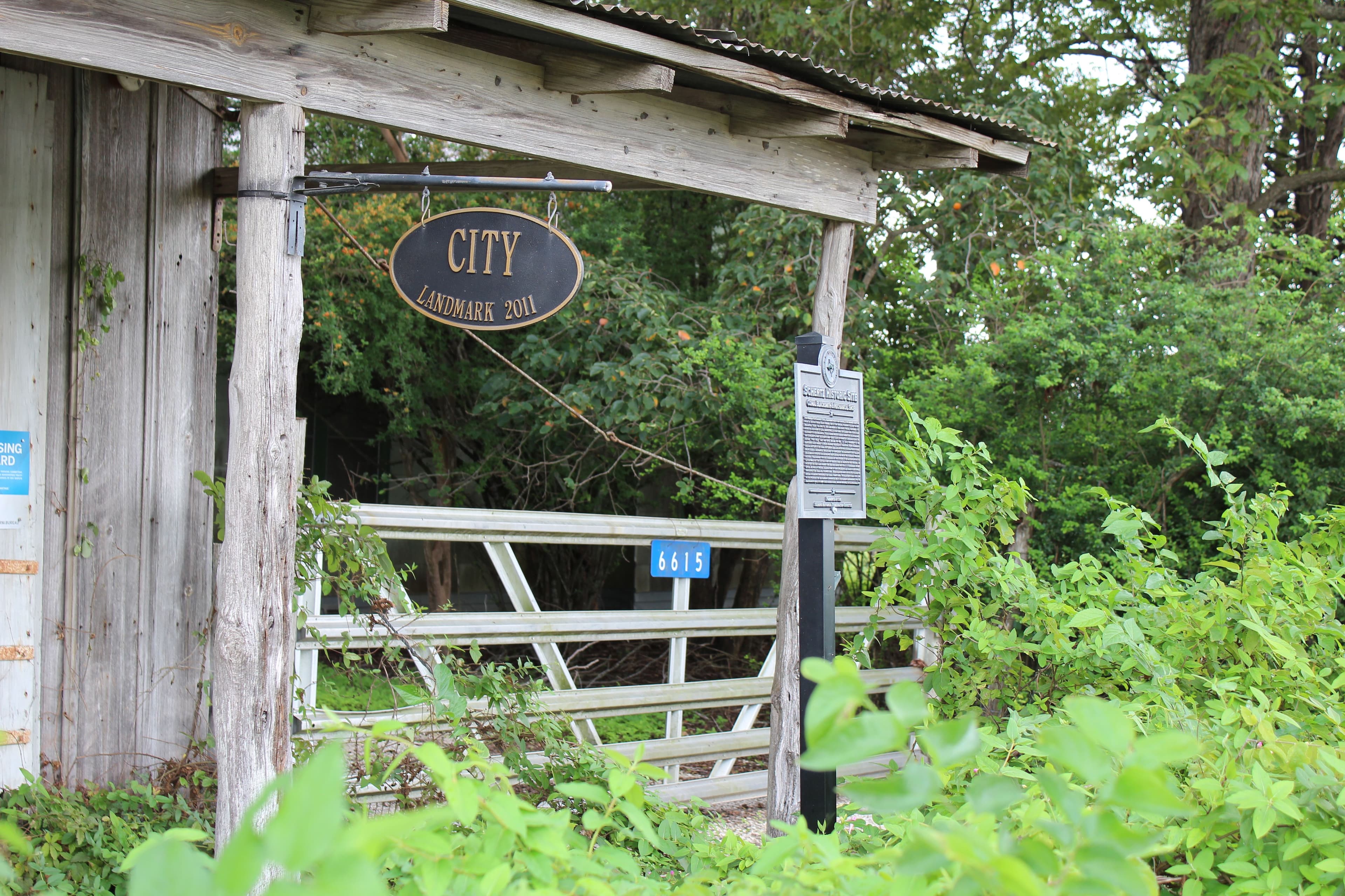 The corner of a weathered grey timber shed with a corrugated metal roof. An oval black-and-gold plaque reading 'CITY LANDMARK 2011' hangs from an iron bracket on the corner post, and a small cast marker on a black post stands to the right. A galvanised farm gate closes the gap between them, and dense green shrubs and trees fill the rest of the frame.