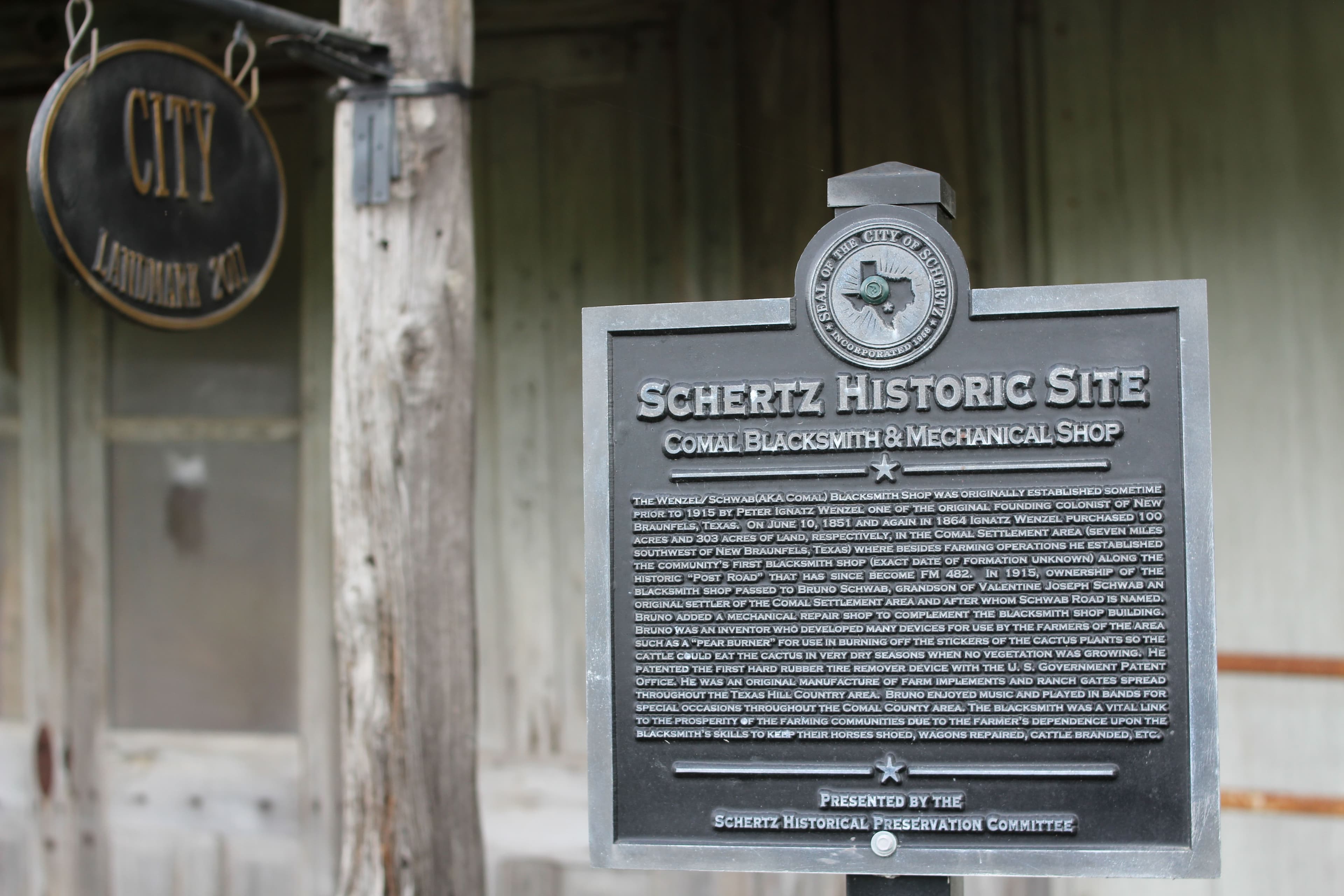 A dark cast-metal plaque on a post, headed by a round city seal with an outline of Texas, reading 'Schertz Historic Site — Comal Blacksmith & Mechanical Shop' above a dense paragraph of raised text. Behind it, out of focus, are the weathered grey boards, corner post and plank door of an old timber shop, with an oval landmark plaque hanging at the left.