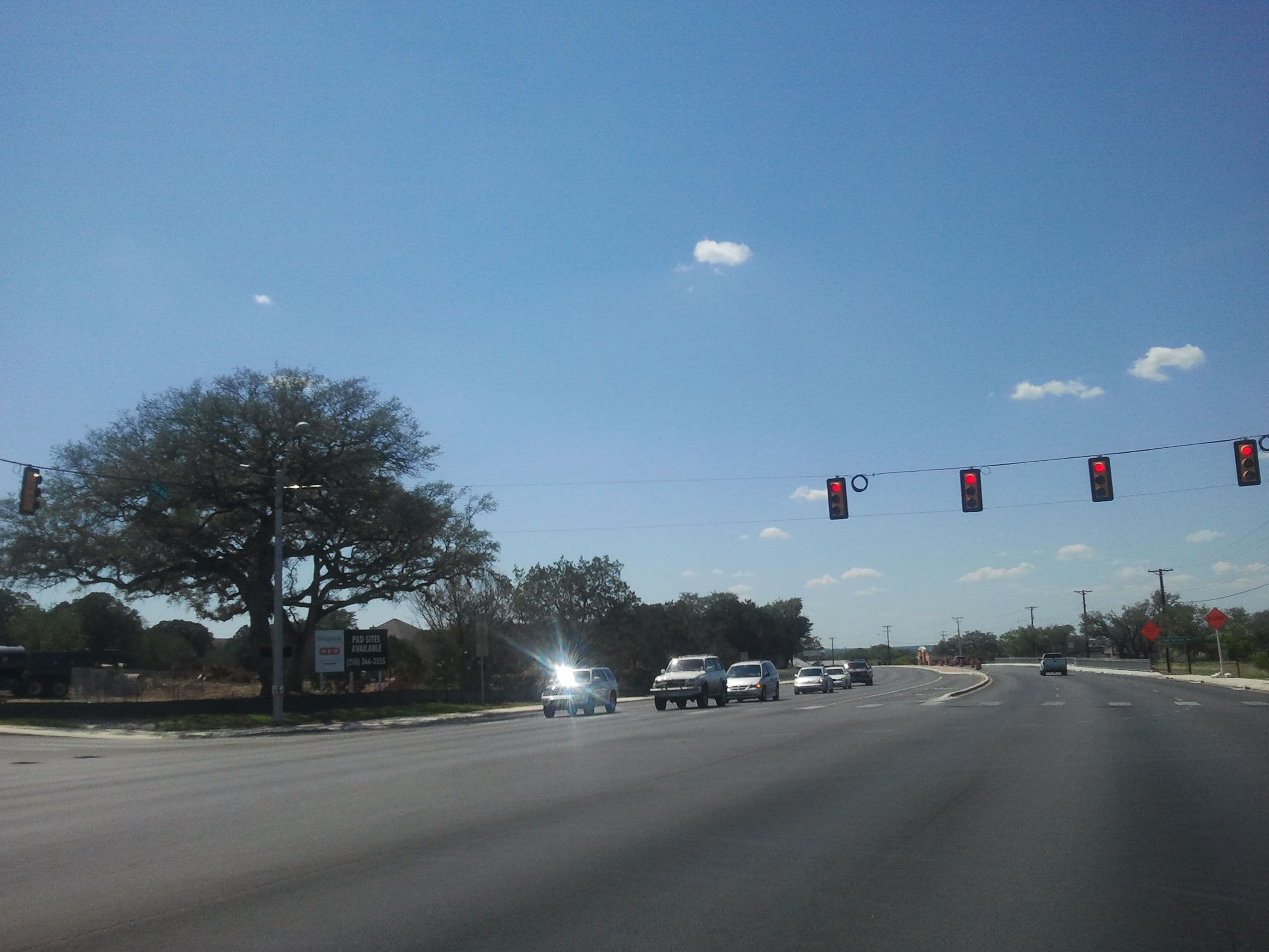 A wide multi-lane highway seen from a stopped vehicle. Traffic signals hung from a span wire show red, oncoming cars and pickups wait with headlights on, and the road curves right past a raised median under a clear blue sky.