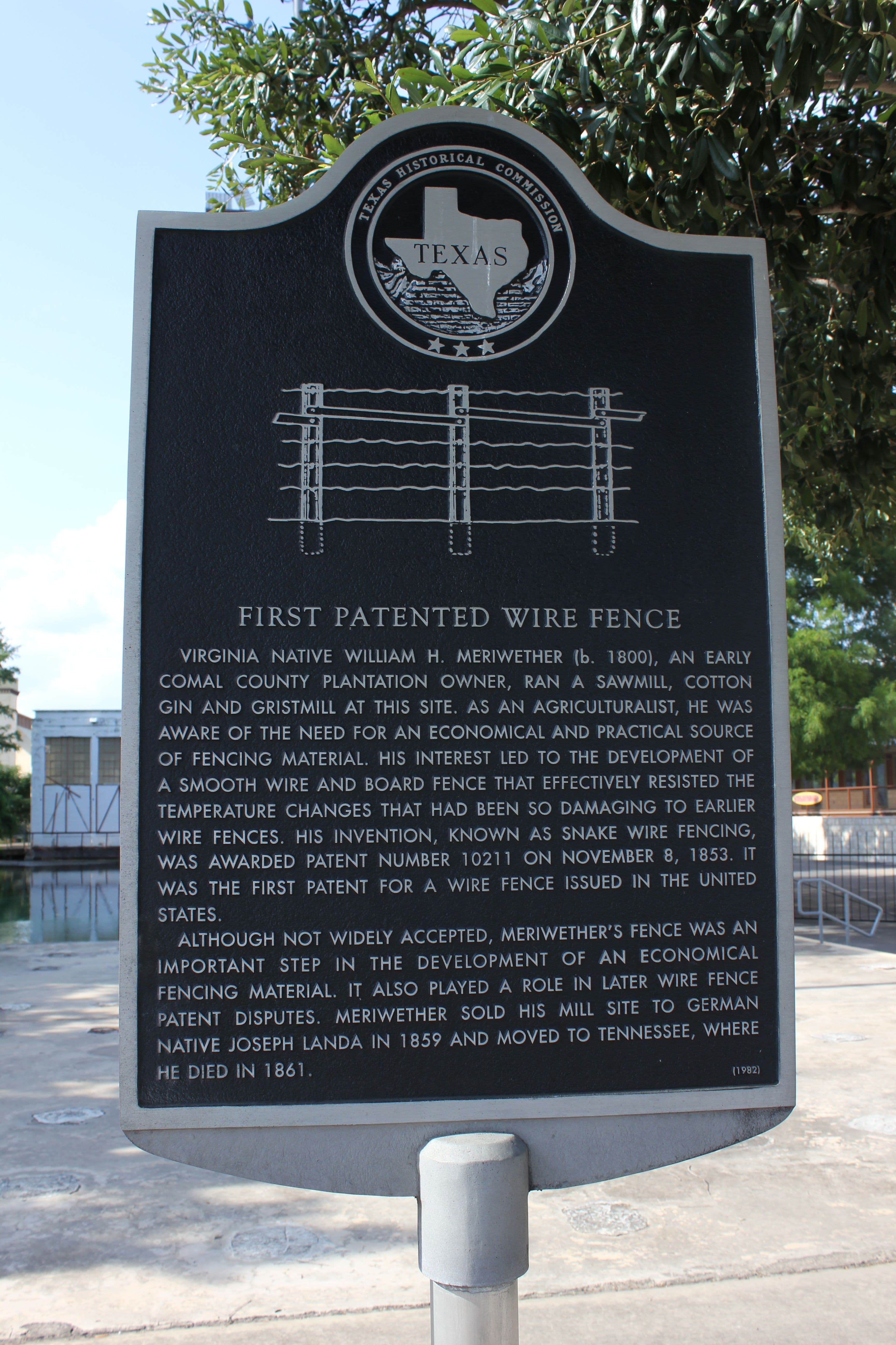 A dark cast-metal historical marker on a grey post, with the Texas seal and a line drawing of a wire fence above several paragraphs of raised text. Behind it are a concrete deck, still green water with a handrail, and a white building among trees.