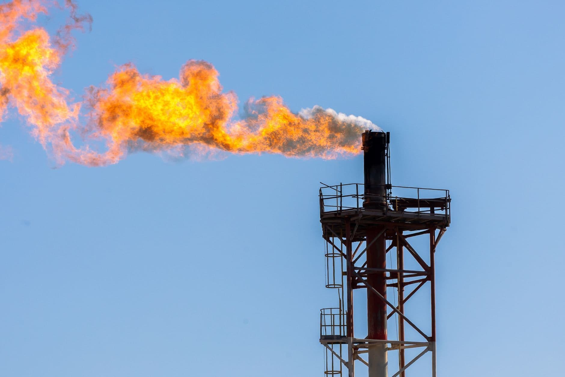 A large orange flame burns from a tall metal industrial stack beneath a blue sky.