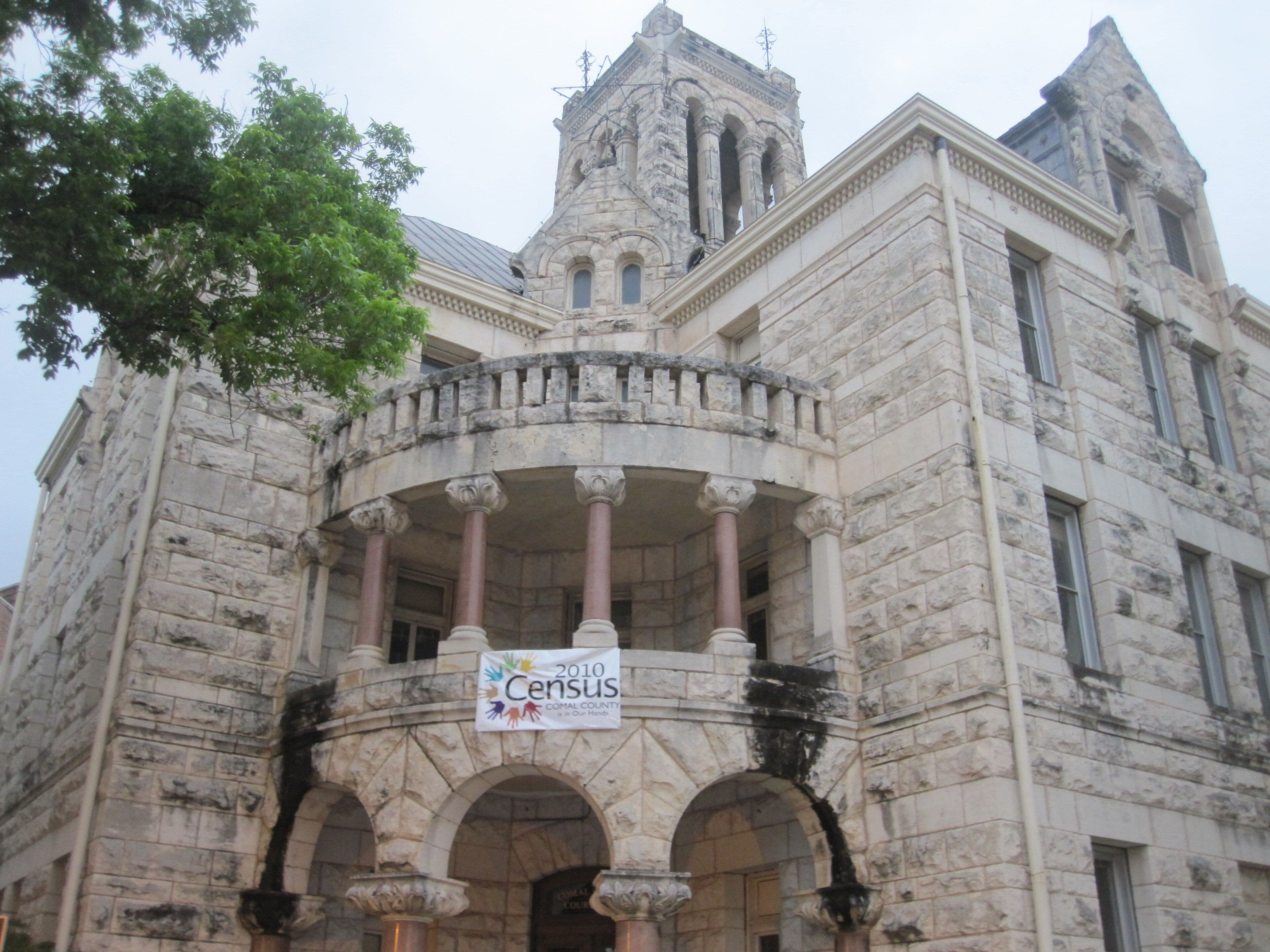 A rusticated pale limestone courthouse photographed from below. A curved stone balcony on pink granite columns sits above a triple-arched entrance, with a square tower behind and a printed banner hung across the balustrade.