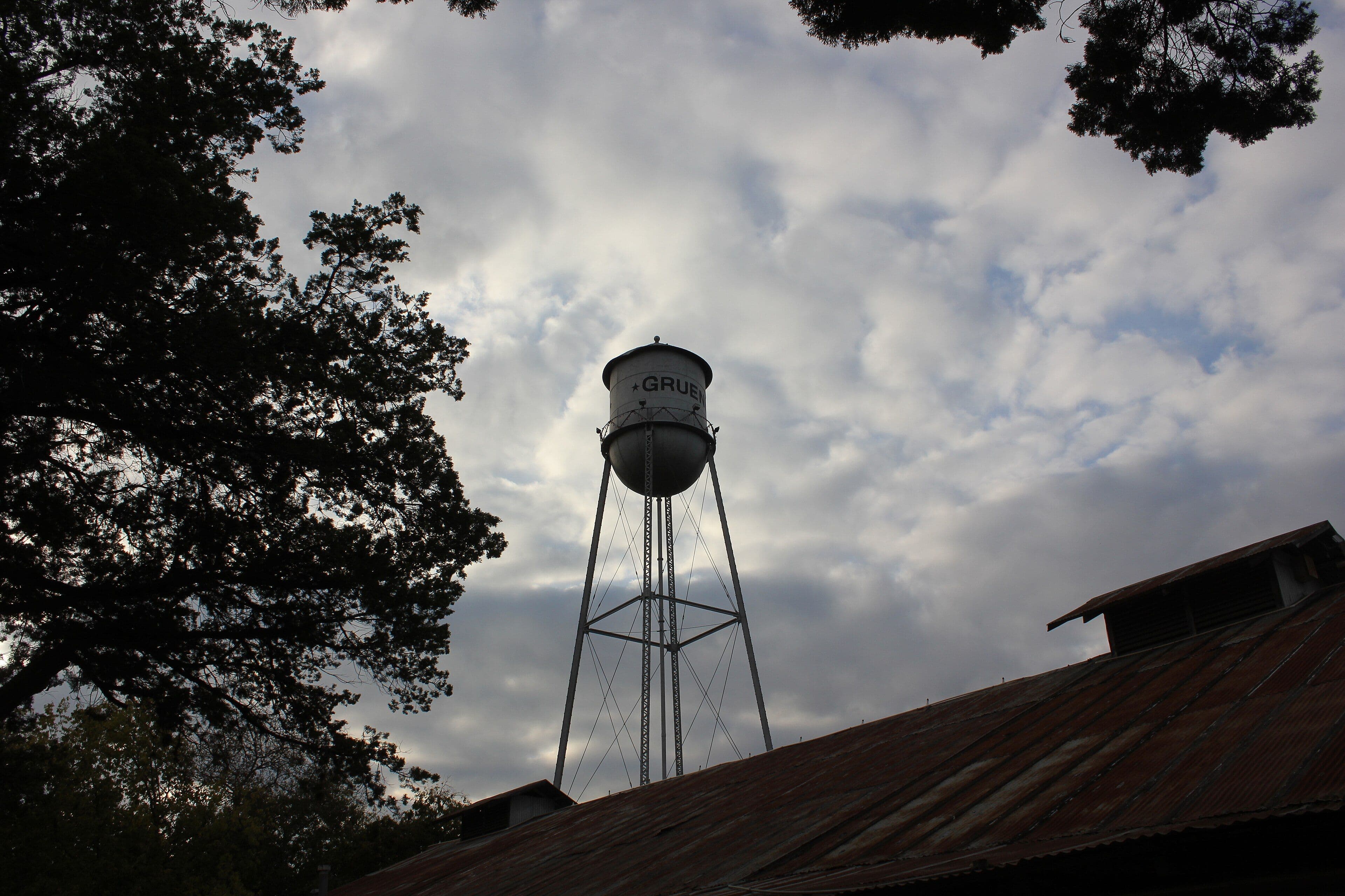 A tall steel water tower with a rounded tank and pointed roof stands in silhouette against a sky of broken grey cloud. Painted lettering and a star are visible on the tank, and the rusted metal roof of a building fills the lower corner.