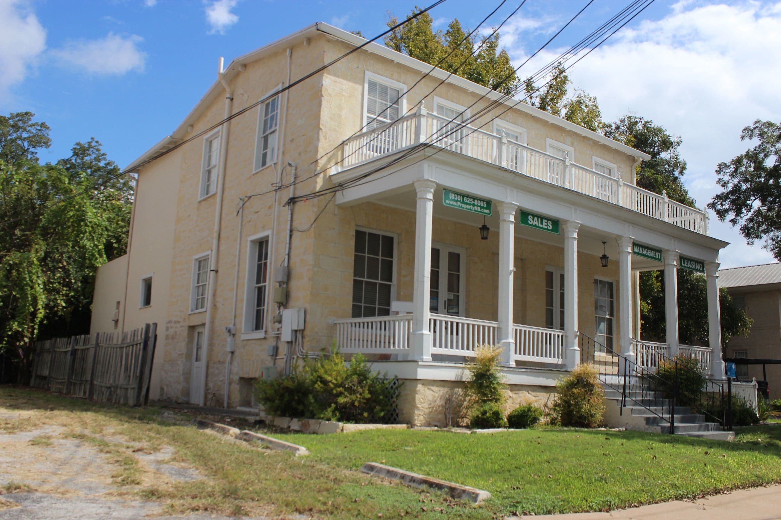 A two-storey cream limestone building seen from the corner of a lawn, with a white timber porch on turned columns, a balustraded gallery above, green business signs on the porch beam, and utility lines crossing overhead.